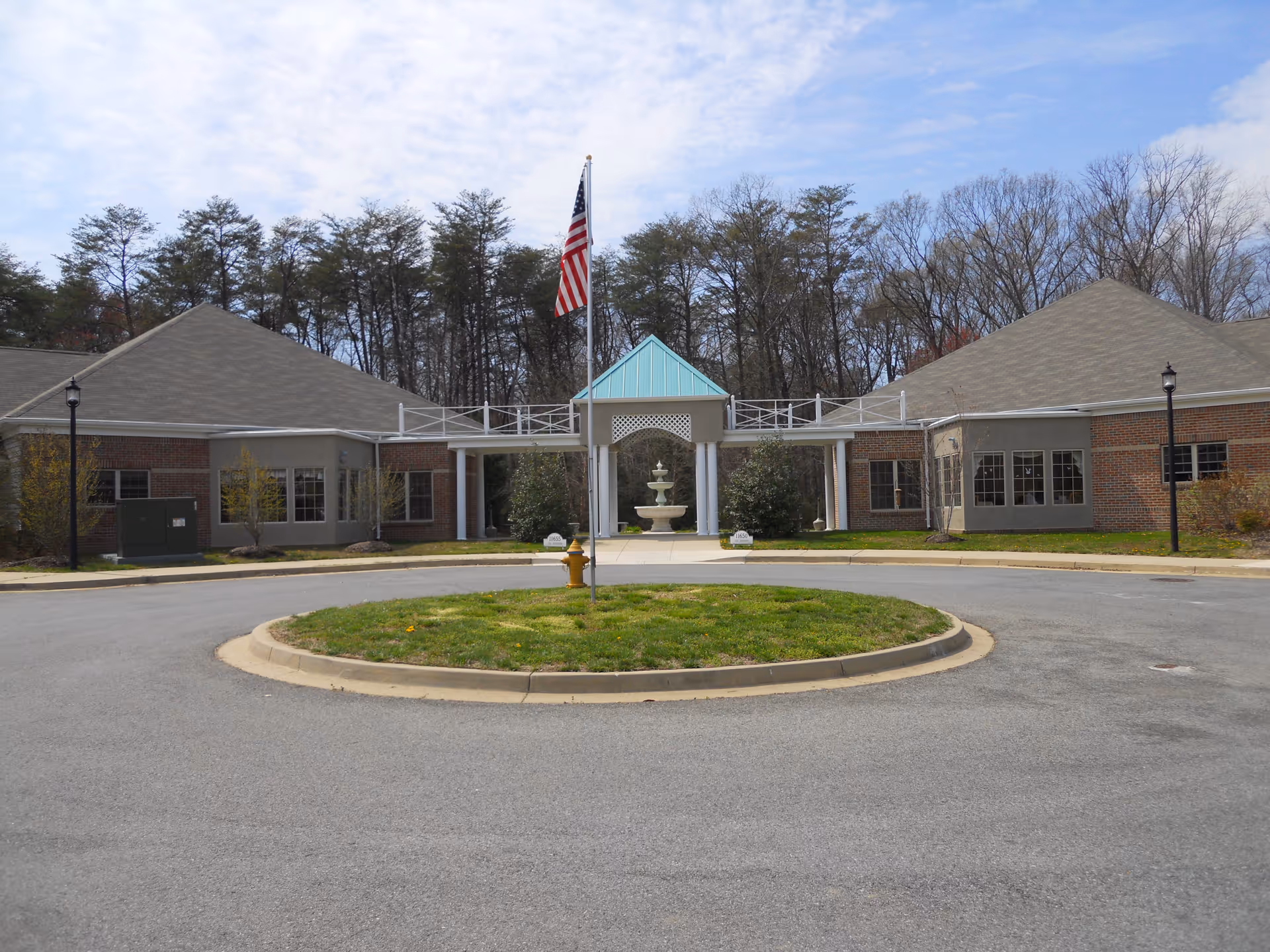 Front exterior view of Fenwick Landing Senior Care Community showing a circular driveway with a grassy roundabout and a fire hydrant in the center. The building has a brick facade with large windows and a covered entrance with white columns and a blue peaked roof. An American flag is flying on a flagpole in the middle of the roundabout. Trees are visible in the background under a partly cloudy sky.