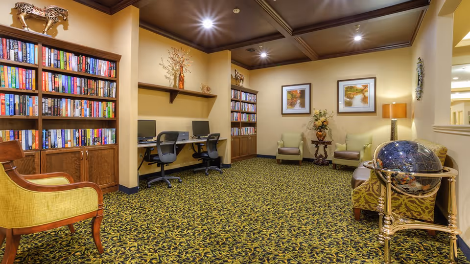 A cozy interior room with patterned carpet, two bookshelves filled with books, two computer desks with chairs, two armchairs with a small table and flower arrangement between them, framed landscape pictures on the wall, and a decorative globe on a stand.
