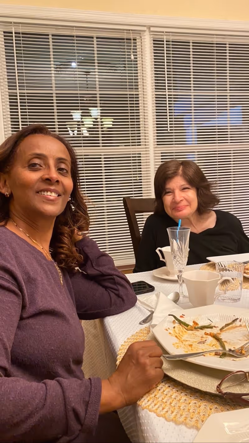 Two women seated at a dining table with finished plates, glasses, and a window with closed blinds behind them.