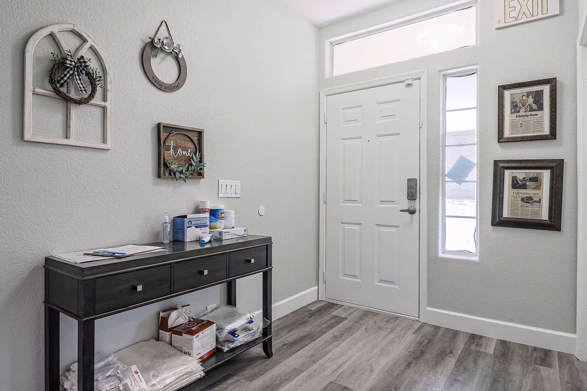 Entryway with a white front door, narrow sidelight window, a dark console table stocked with supplies, and decorative wall hangings.