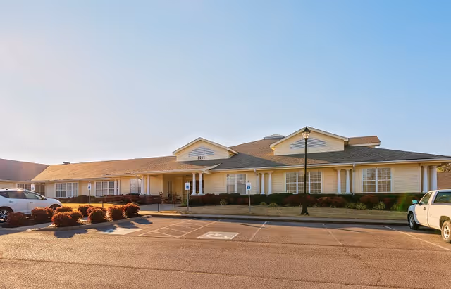 Exterior view of a single-story senior living facility building with a beige facade, multiple windows, a covered entrance, and a parking lot with handicap parking spaces in front. There are some bushes and a street lamp near the entrance.