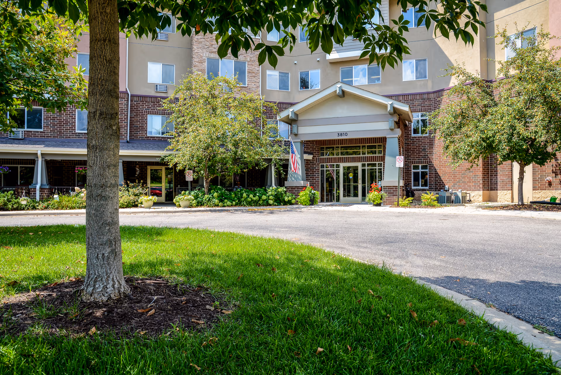 Front entrance of a senior living building with a covered porte-cochere, driveway, lawn, and trees.