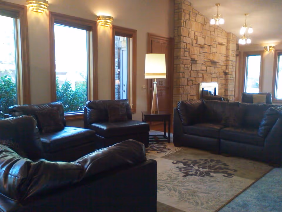 A cozy living room with dark leather sofas and armchairs arranged around a patterned area rug. The room features large windows with wooden frames letting in natural light, a stone fireplace, a floor lamp, and wall-mounted lights.