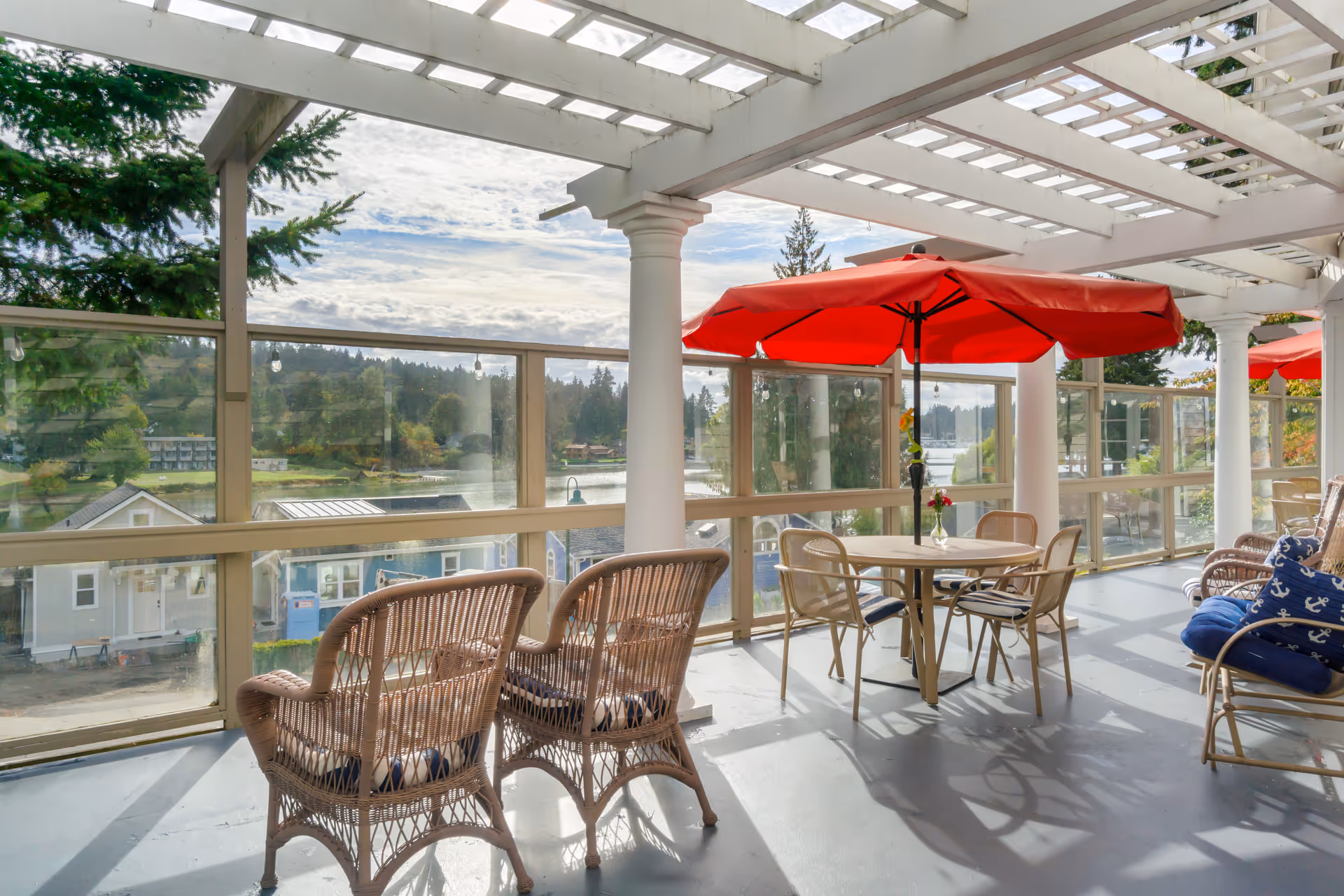 Covered outdoor terrace with wicker chairs, tables and red umbrellas overlooking a waterfront through a glass railing.