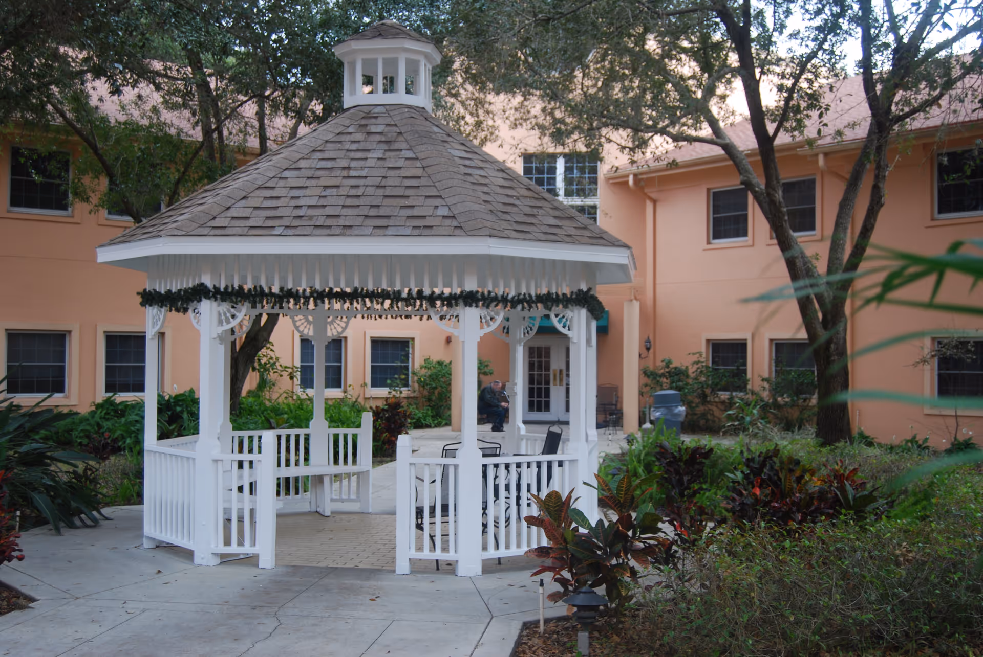 White wooden gazebo with a shingled roof decorated with garland, situated in a garden area with trees and shrubs, in front of a peach-colored building with multiple windows.
