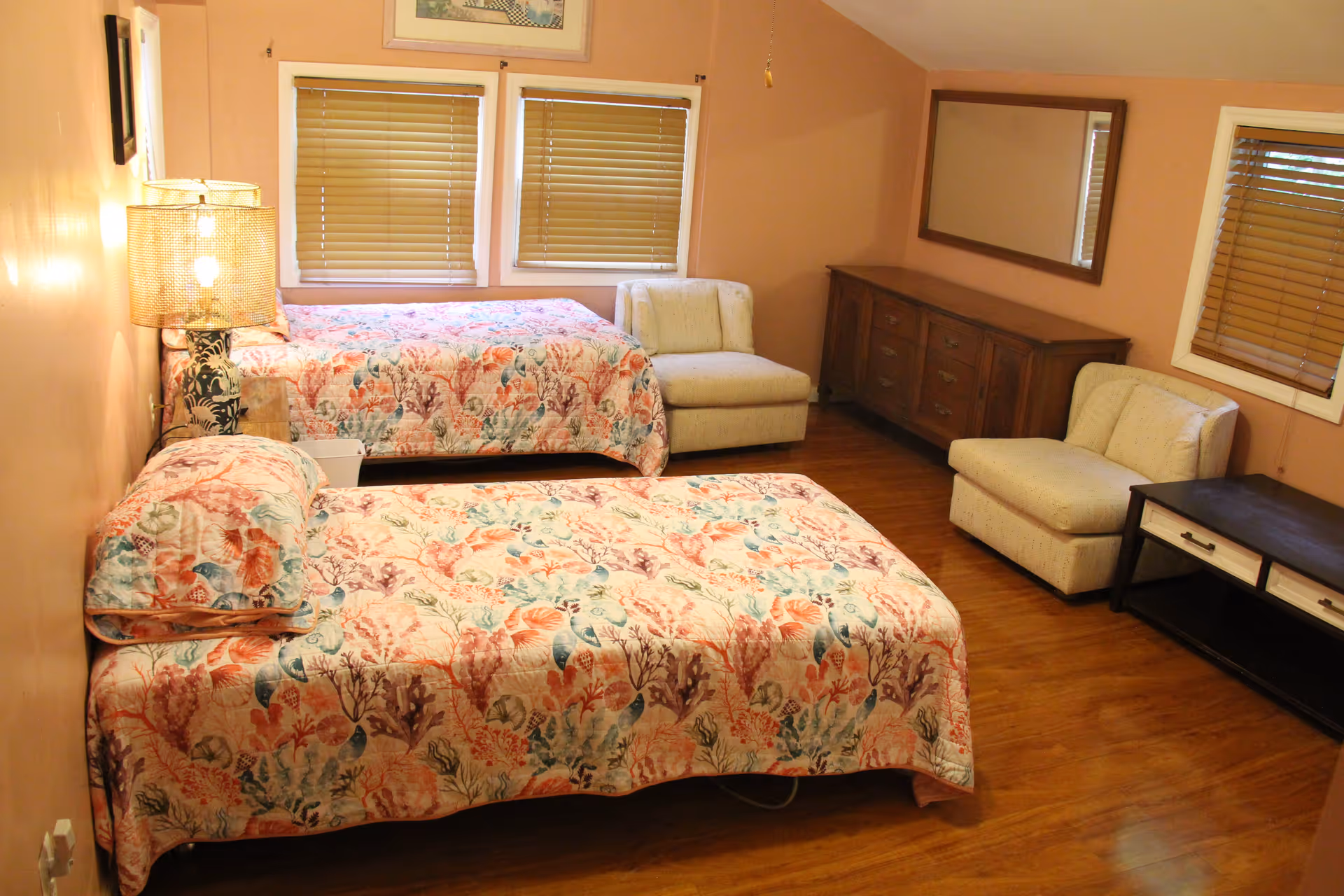 Warm-toned bedroom with two twin beds in floral bedspreads, two chairs, a dresser and window blinds.
