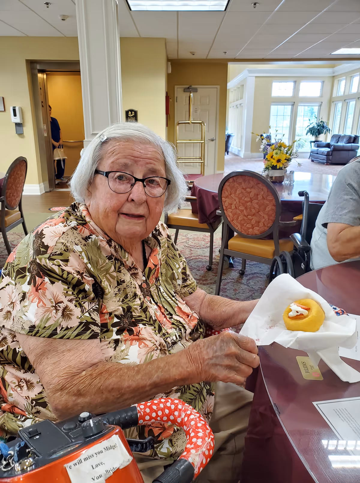 An elderly woman with white hair and glasses sitting at a table in a communal dining area, holding a napkin with a small dessert. She is wearing a floral patterned shirt and is seated next to a walker with a red and white polka dot cover. The background shows chairs, tables, and a bright room with large windows and plants.