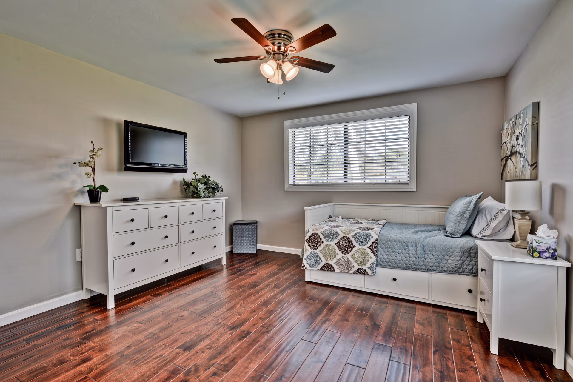 A bright bedroom with wooden flooring, a white daybed with blue bedding and a patterned throw blanket, a white nightstand with a lamp and tissue box, a white dresser with plants on top, a wall-mounted flat screen TV, a window with blinds, and a ceiling fan with lights.