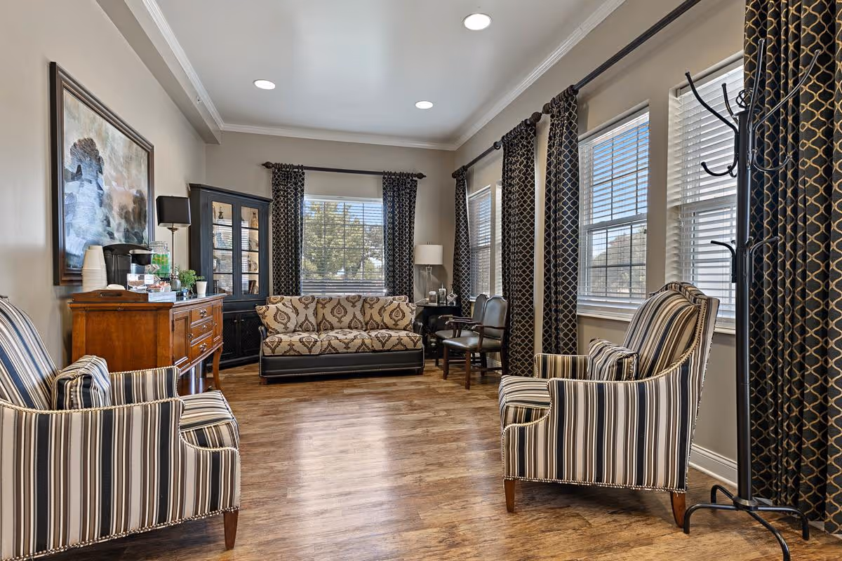 A well-lit living room with wooden flooring, featuring a patterned sofa against the back wall, two striped armchairs in the foreground, a wooden sideboard with a coffee maker and cups, a black display cabinet, and several windows with patterned curtains allowing natural light to enter.
