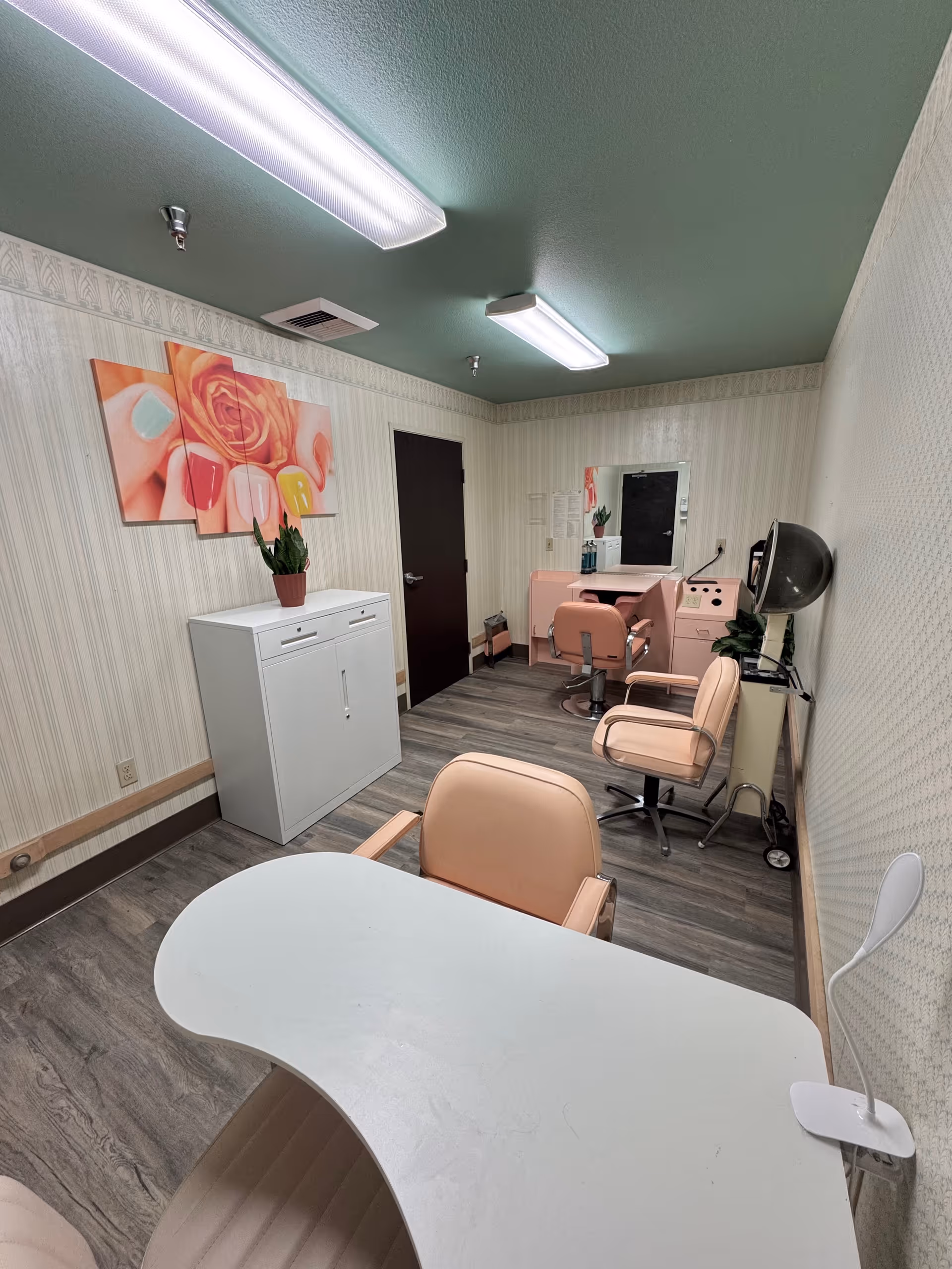 Interior view of a small salon or beauty room with a white curved table and beige chair in the foreground, two beige salon chairs, a pink vanity with a mirror, a hair dryer hood, a white cabinet with a potted plant on top, and a wall art featuring a close-up of painted nails and a rose.