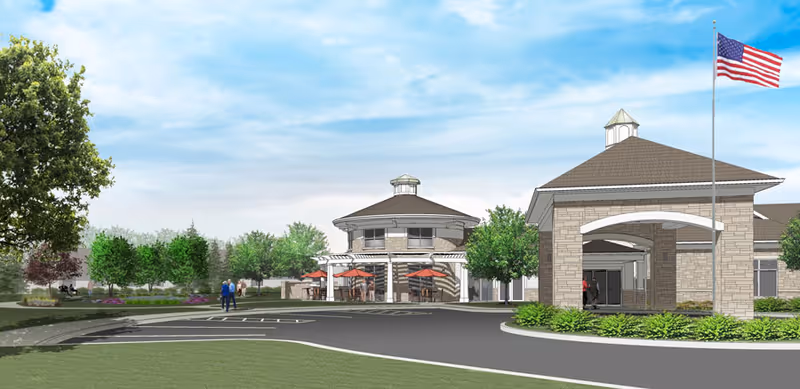 Exterior view of Vista Springs Timber Ridge Village showing a driveway leading to a covered entrance with an American flag on a flagpole. The building has a stone facade and a peaked roof. In the background, there is a round building with outdoor seating shaded by red umbrellas, surrounded by trees and greenery under a partly cloudy sky.