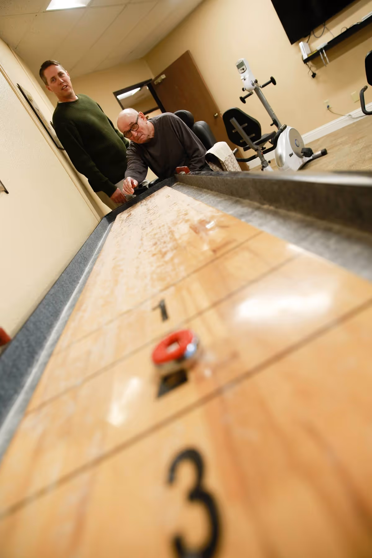 Two men playing shuffleboard indoors at Gladding Ridge Assisted Living and Memory Care. One man is seated and focused on the game, while the other stands nearby watching. Exercise equipment and a wall-mounted TV are visible in the background.