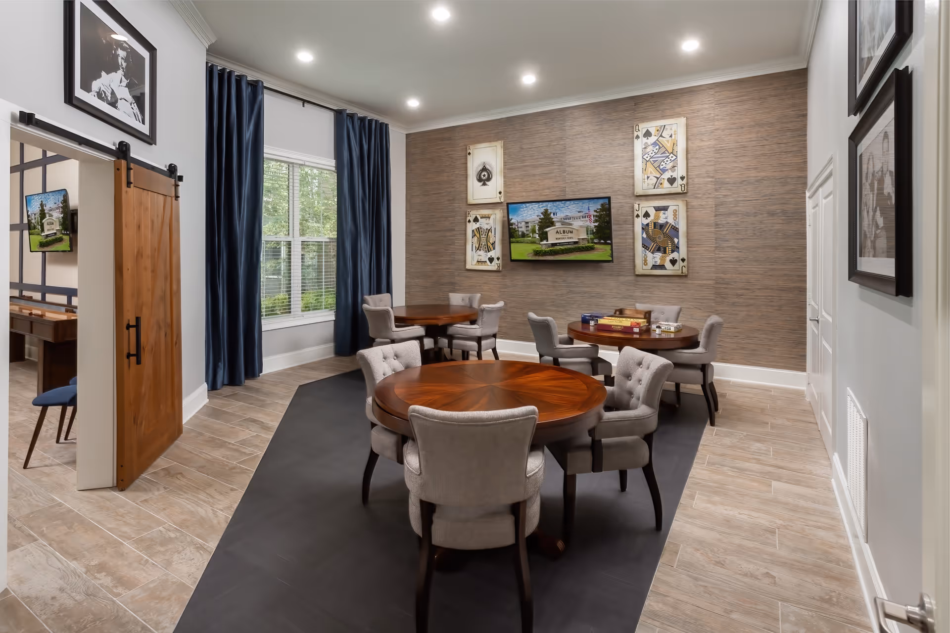 A well-lit game room with three round wooden tables, each surrounded by four cushioned chairs. The walls are decorated with large playing card artwork and a flat-screen TV displaying an image of the Album Monterey Pointe sign. There is a window with blue curtains letting in natural light, and a wooden sliding barn door partially open to an adjacent room.