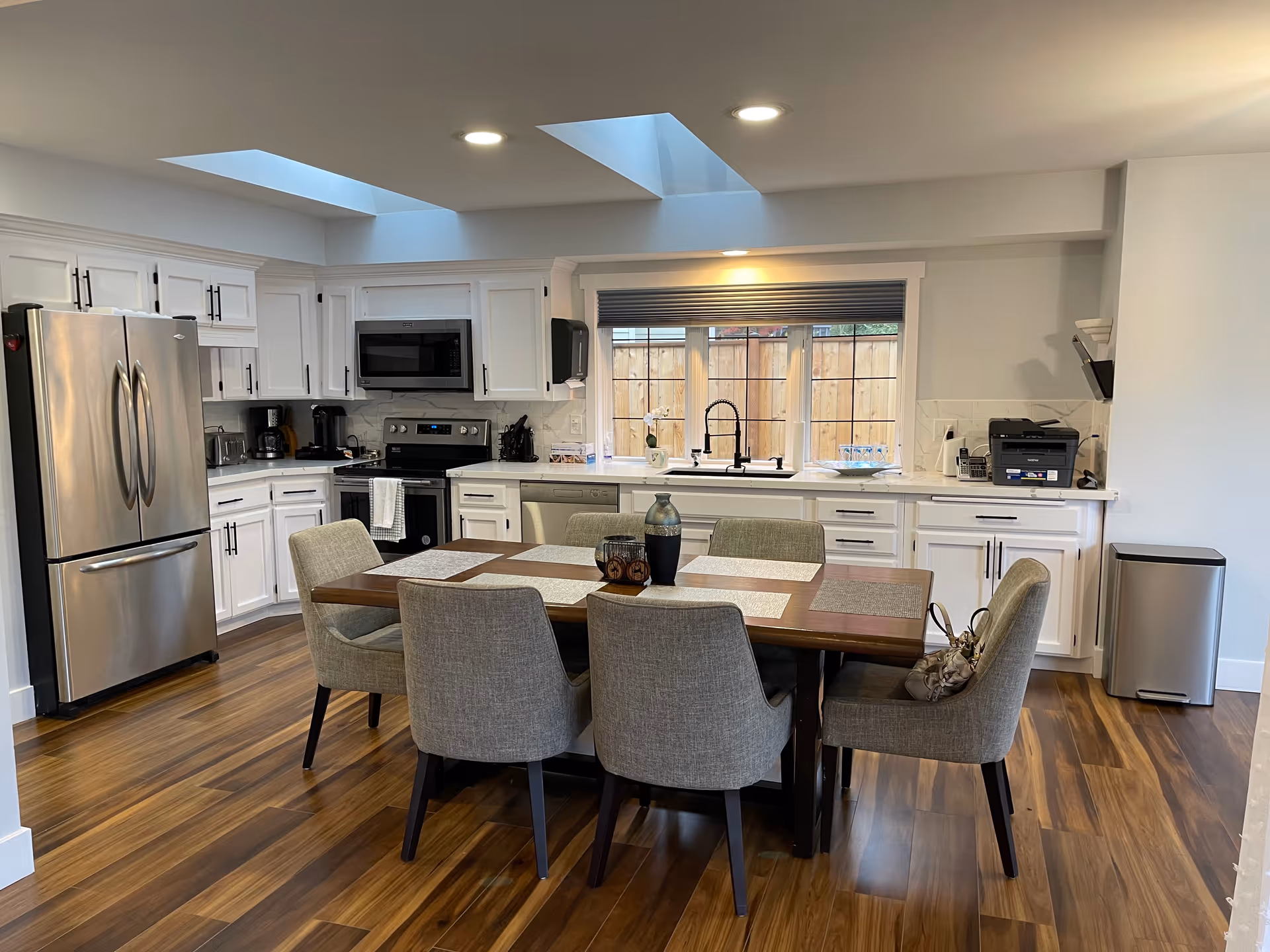 A modern kitchen with white cabinets, stainless steel refrigerator, microwave, stove, and dishwasher. A wooden dining table with six gray upholstered chairs is centered in the room. The kitchen has wooden flooring, two skylights, and a window with a view of a wooden fence. Various kitchen appliances and items are on the countertops.