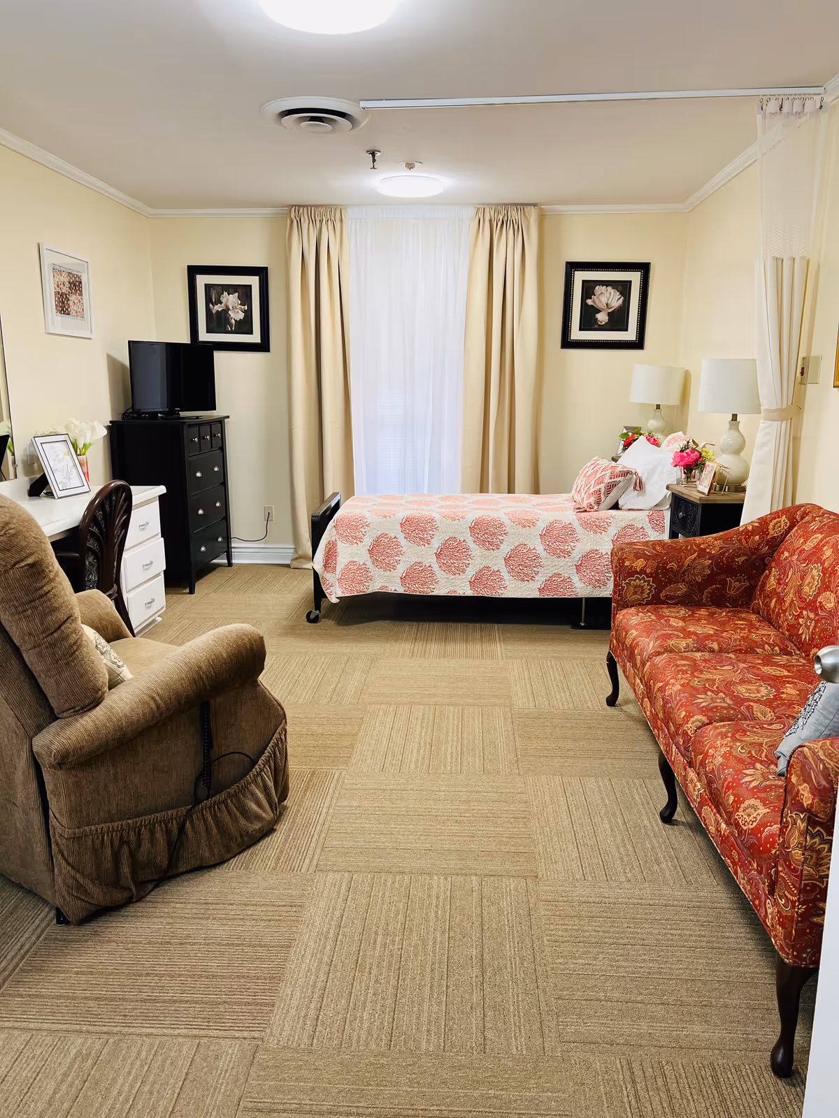 A cozy senior living bedroom with a bed, red patterned sofa, brown recliner, dresser with TV, and cream curtains.