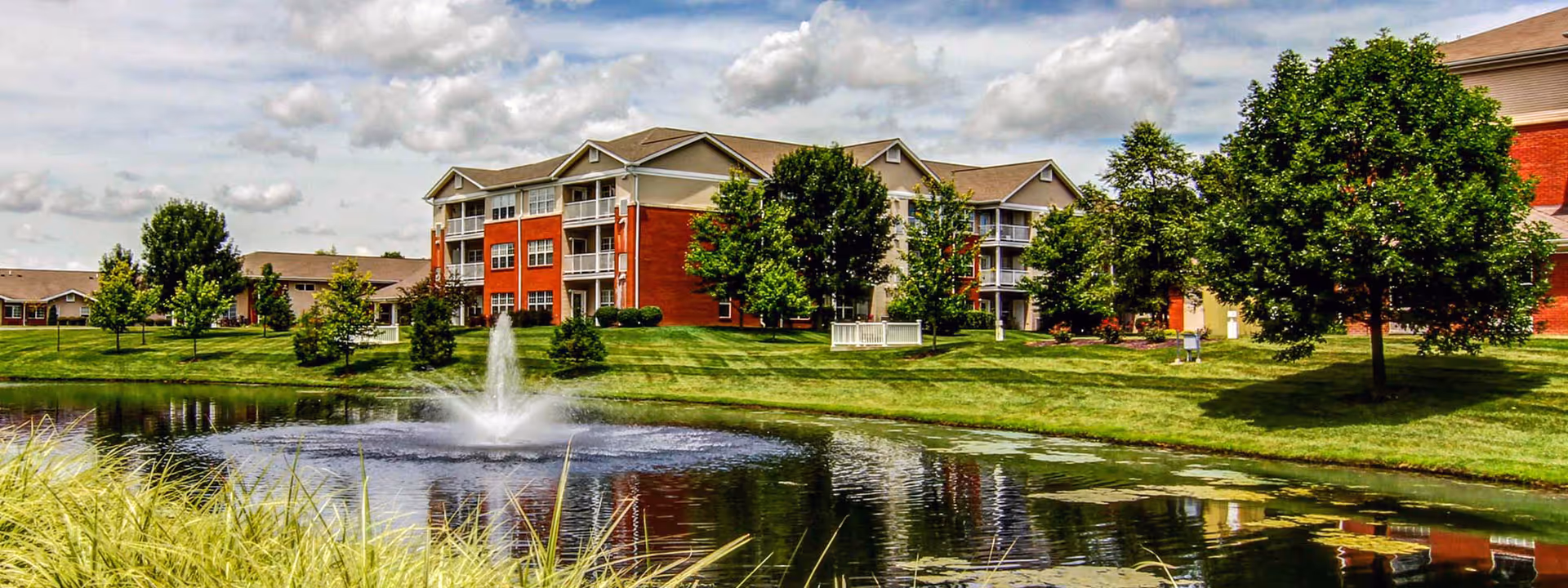 A scenic view of EverTrue Meridian Village showing a large pond with a water fountain in the center, surrounded by well-maintained green lawns and trees. In the background, there are multi-story residential buildings with red brick and beige siding under a partly cloudy sky.