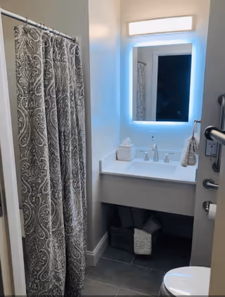 A small bathroom featuring a patterned shower curtain on the left, a white sink with a modern faucet and a backlit rectangular mirror above it. Below the sink are two baskets, one containing towels. A toilet is partially visible on the right side, along with a grab bar mounted on the wall.