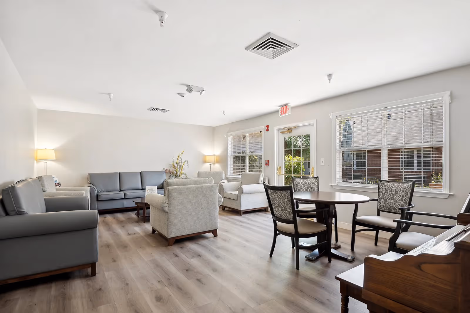 A bright and spacious living room area in a senior living facility with multiple gray sofas and armchairs arranged around a coffee table. There is a round table with four patterned chairs near large windows that let in natural light. The room has light-colored walls, wood flooring, two lamps, and a piano on the right side. An exit door is visible in the background.