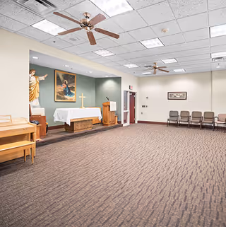 A spacious room with carpeted floor and ceiling fans, featuring a small altar with a white cloth, a cross, a statue of Jesus, a painting, and a lectern. Several chairs are lined up against the far wall.