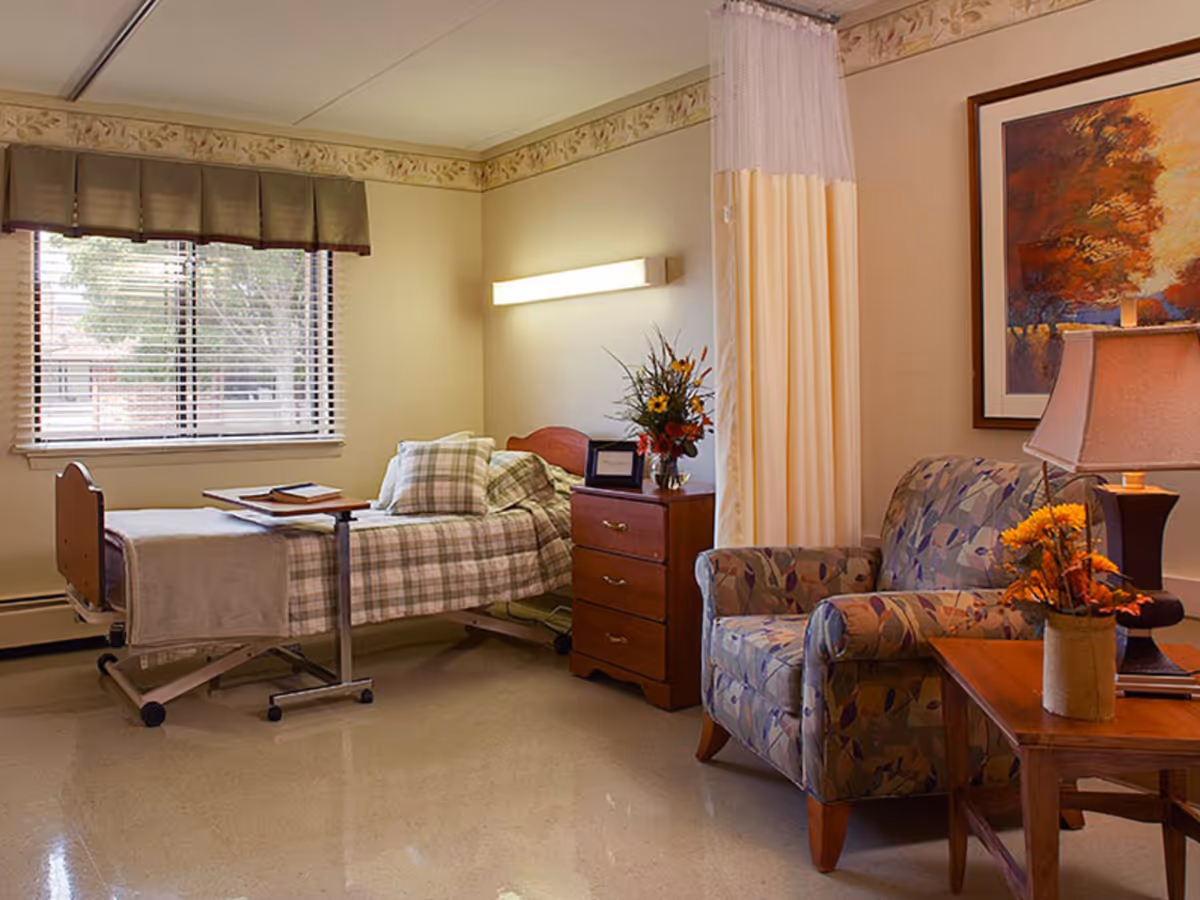 A cozy senior living bedroom with a single bed covered in plaid bedding, a wooden nightstand with a vase of flowers and a framed picture, a patterned armchair, a wooden side table with a lamp and a flower arrangement, a window with blinds and a valance, and a framed autumn-themed painting on the wall.