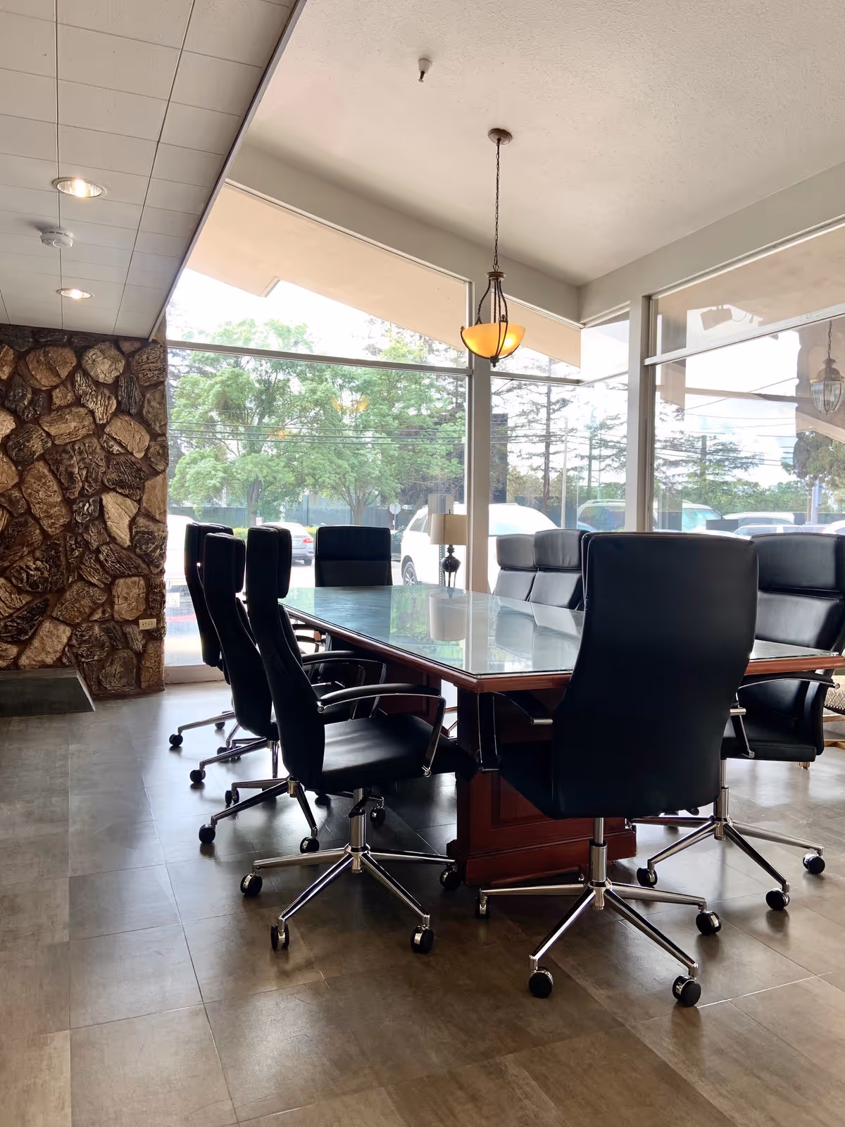 A conference room with a large rectangular glass-top table surrounded by black office chairs on wheels. The room has large windows letting in natural light and a stone accent wall on one side. A hanging light fixture is above the table.