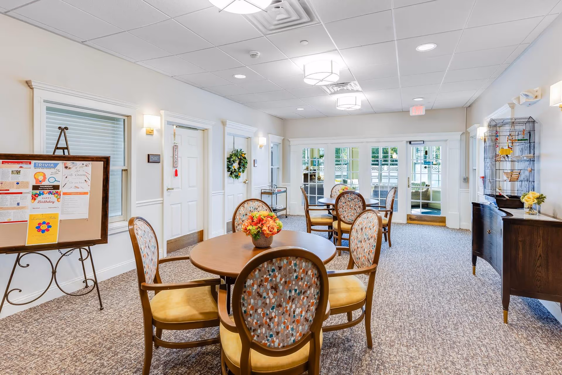 Bright, well-lit communal dining/activity room with round tables, patterned chairs, a bulletin board and glass doors leading outside.