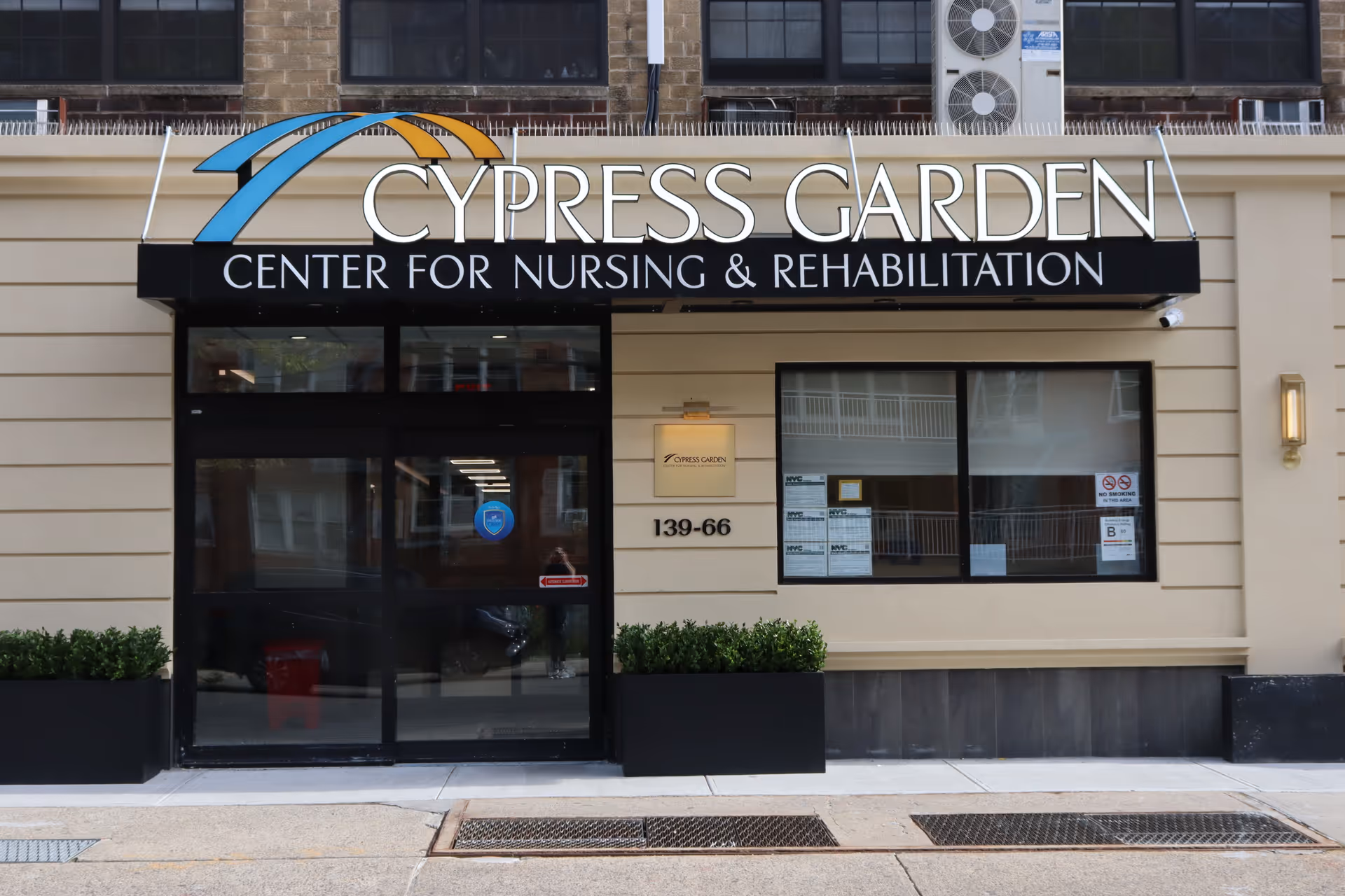 Front entrance of Cypress Garden Center for Nursing & Rehabilitation with a black awning displaying the facility name and address 139-66. The entrance has glass doors and windows, with two black planters containing green shrubs on either side.