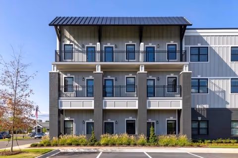 Exterior view of a modern three-story building with balconies on the second and third floors, large windows, and a metal roof. The building is surrounded by landscaping with grass and shrubs, and there is a parking area in front.