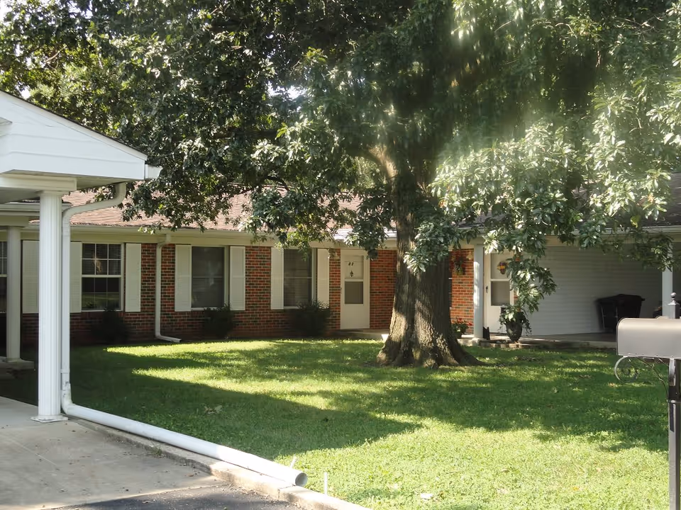 Exterior view of a single-story brick building with white window shutters and a white door, partially shaded by a large tree in the front yard with green grass. A white gutter and downspout are visible along the edge of the roof, and a black mailbox is in the foreground.