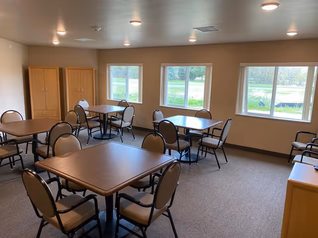 A bright room with multiple square wooden tables and beige cushioned chairs arranged around them. The room has three large windows showing a green outdoor view, beige walls, and carpeted flooring. There are wooden cabinets against one wall.