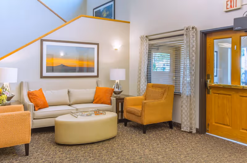 A cozy living room area with a beige sofa adorned with orange pillows, two orange armchairs, a round beige ottoman, two side tables with lamps, a large framed sunset photograph on the wall, a window with patterned curtains, and a wooden door with a glass panel and an exit sign above it.