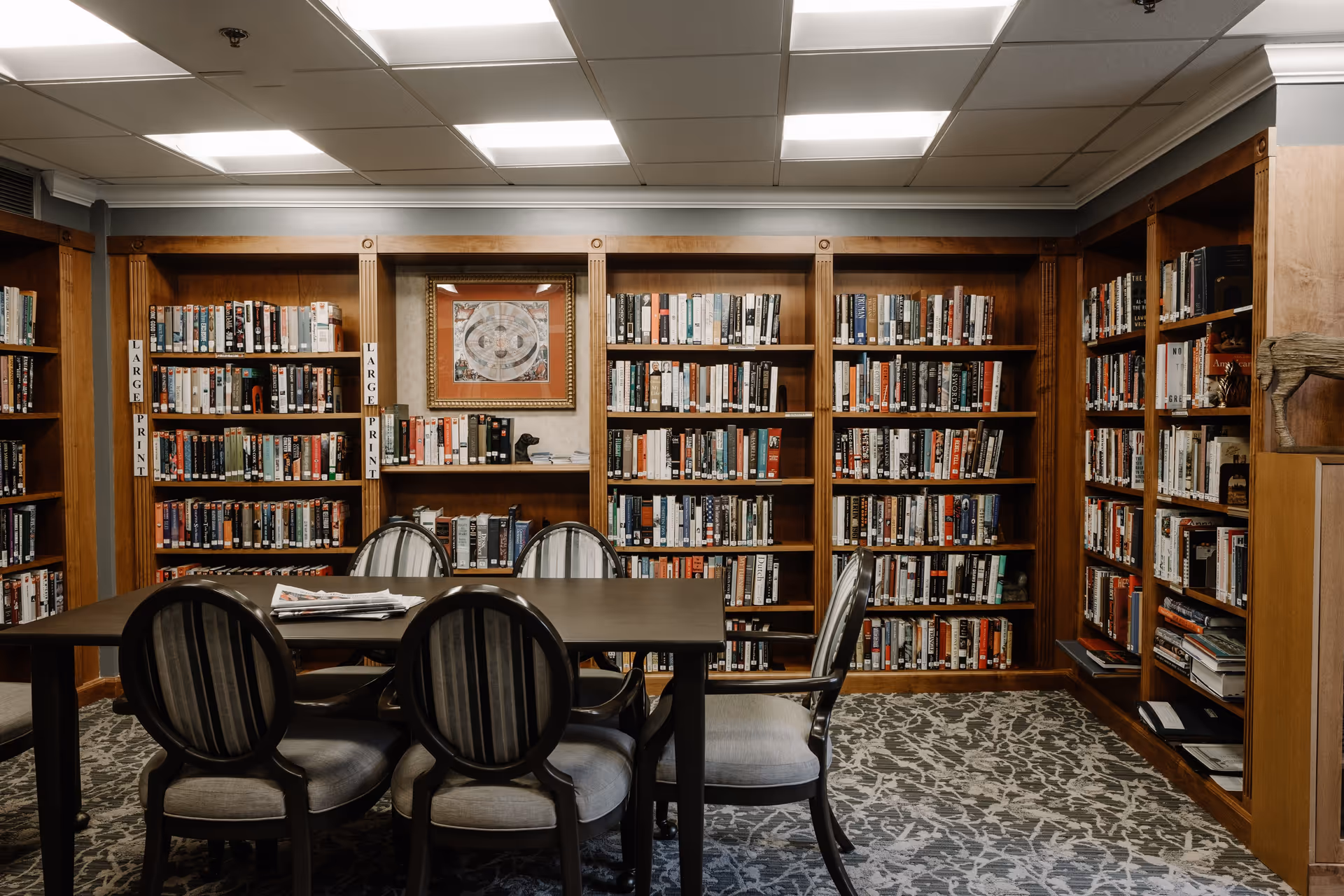 A cozy library room with wooden bookshelves filled with books, a table with four cushioned chairs around it, and a framed artwork on the wall. The shelves have labels indicating 'LARGE PRINT' books. The room has a patterned carpet and ceiling lights.