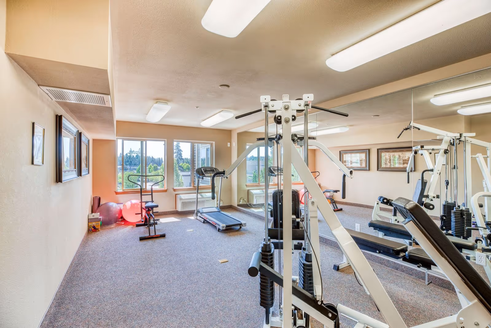 Well-lit interior fitness room with weight machines, a treadmill, stationary bike, exercise balls and mirrored wall.