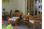 Outdoor patio area with wicker furniture including two chairs with striped cushions, a small table with a potted plant, and a round table with chairs in the background. The patio is adjacent to a building with sliding glass doors and light-colored siding.