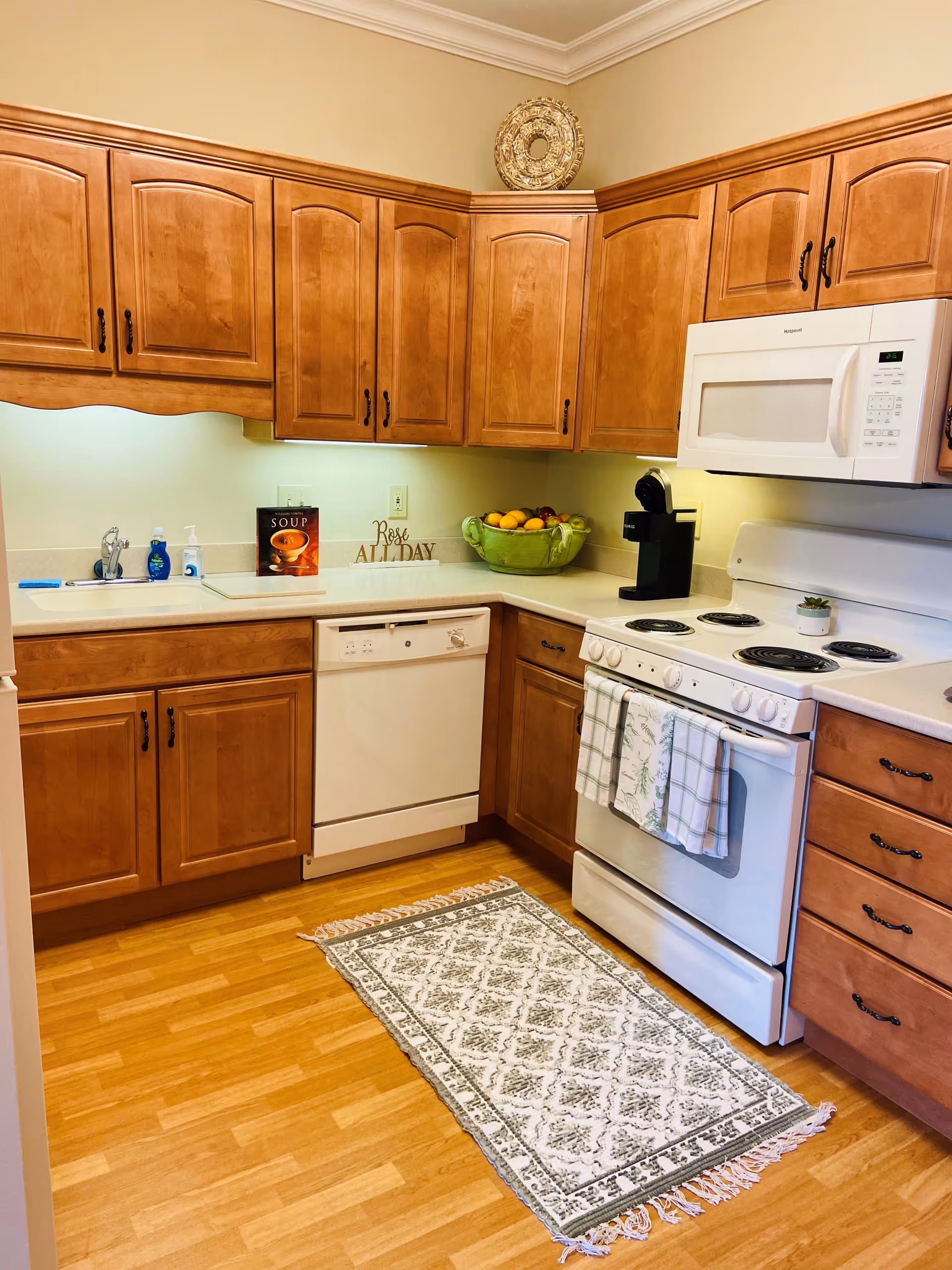 A clean kitchen with wooden cabinets, a white dishwasher, stove, and microwave. There is a patterned rug on the wooden floor, a bowl of fruit on the counter, a coffee maker, and decorative items including a sign that says 'Rose ALL DAY' and a book titled 'SOUP'.