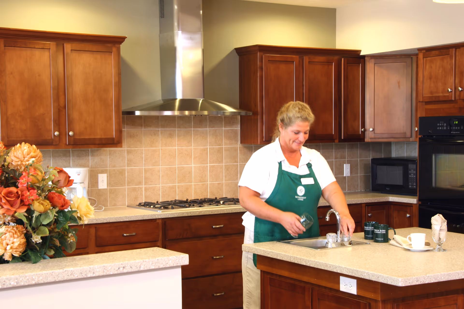 A staff member in a green apron washes a glass at the sink in a kitchen with wooden cabinets and a center island.
