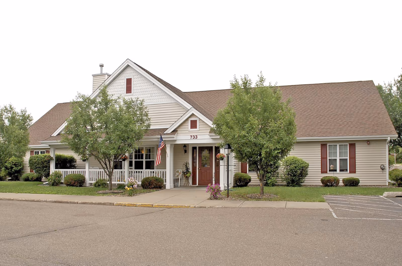 Front exterior of a single-story residential-style building with a covered porch, American flag, and trees in the lawn.