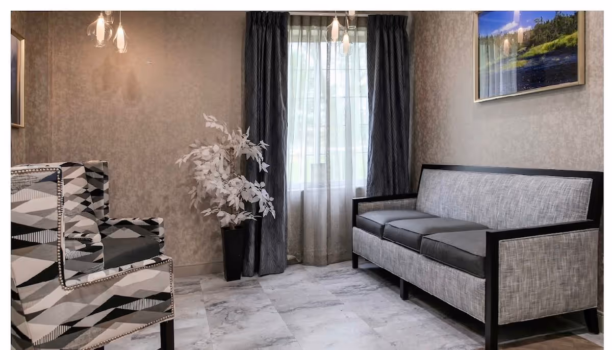 A small sitting area with a patterned armchair on the left and a gray cushioned sofa on the right. Between them is a tall black vase with white decorative branches. Behind the sofa is a window with sheer white curtains and dark drapes. A framed landscape picture hangs on the wall above the sofa. The floor is covered with light-colored tiles.