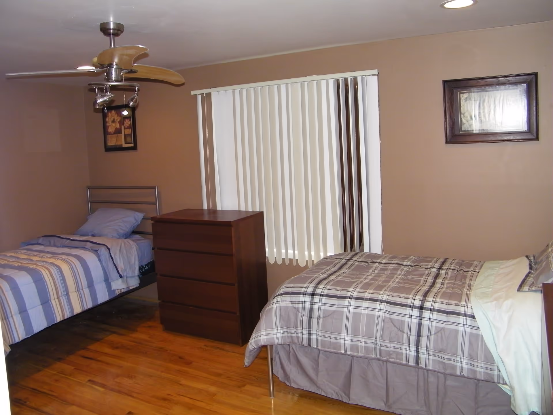 Bedroom with two twin beds, a central chest of drawers, vertical blinds, a ceiling fan and hardwood floor.
