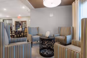 A cozy seating area in a senior living facility with four striped armchairs arranged around a black decorative round table. The room has a neutral color palette with beige walls, a window with blinds, and a ceiling light fixture. In the background, there is a dining area with tables and chairs.