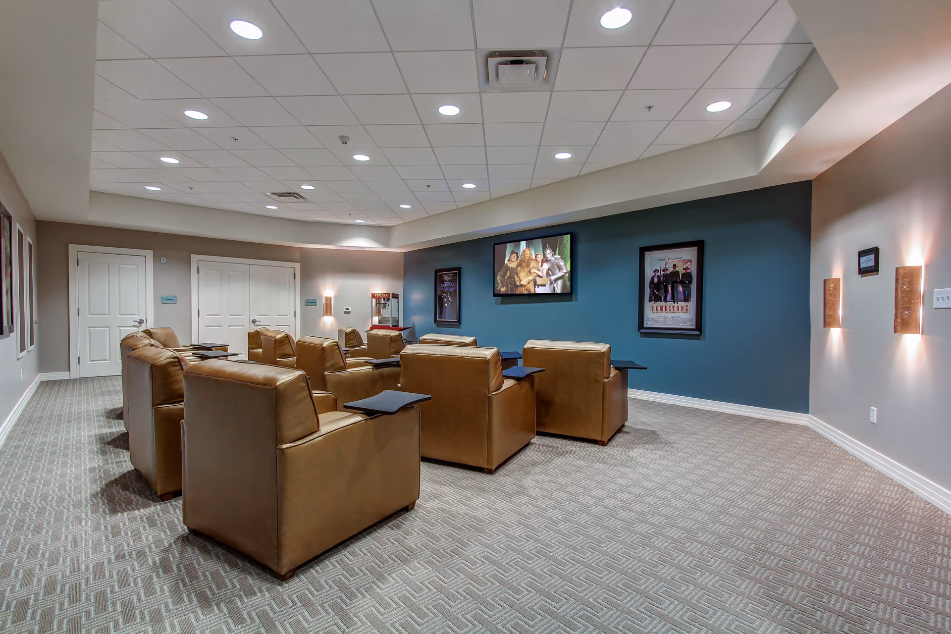 A cozy movie theater room with several brown leather armchairs arranged in rows facing a wall-mounted TV screen showing a scene from The Wizard of Oz. The room has a patterned carpet, soft lighting on the walls, movie posters, and a popcorn machine in the corner.