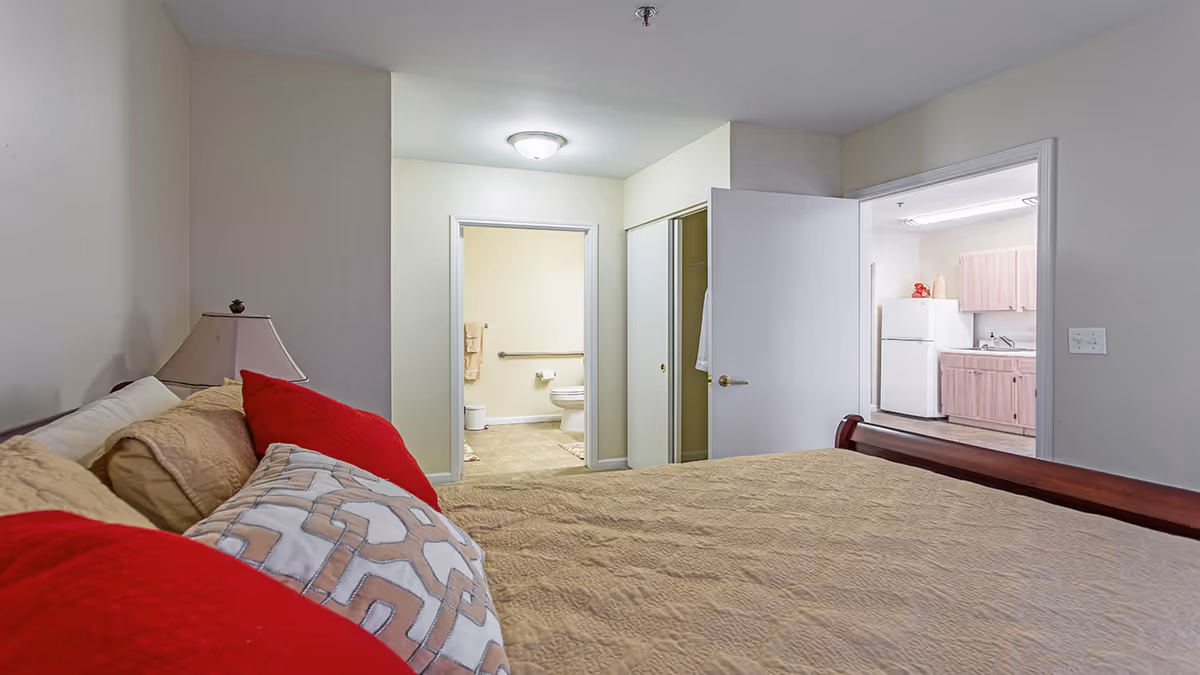 Bedroom featuring a bed with red accent pillows, a bedside lamp, and open doors revealing a bathroom and a kitchenette.