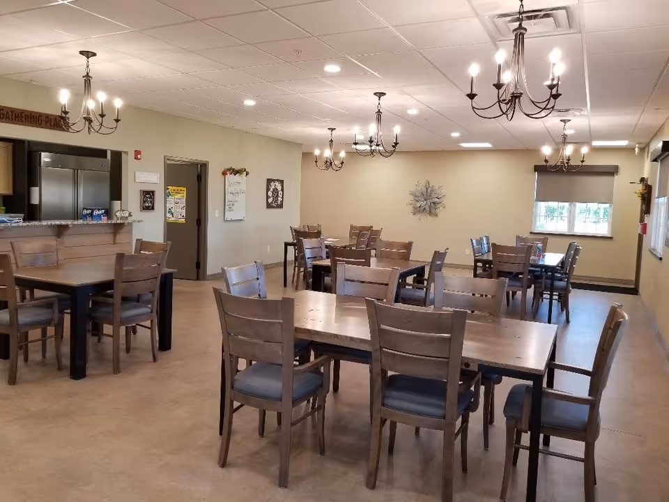 A dining room with multiple wooden tables and chairs arranged neatly. The room has beige walls, a window with a roller shade, and several chandeliers hanging from the ceiling. There is a kitchen area visible in the background with a refrigerator and a counter. The space is well-lit with ceiling lights.