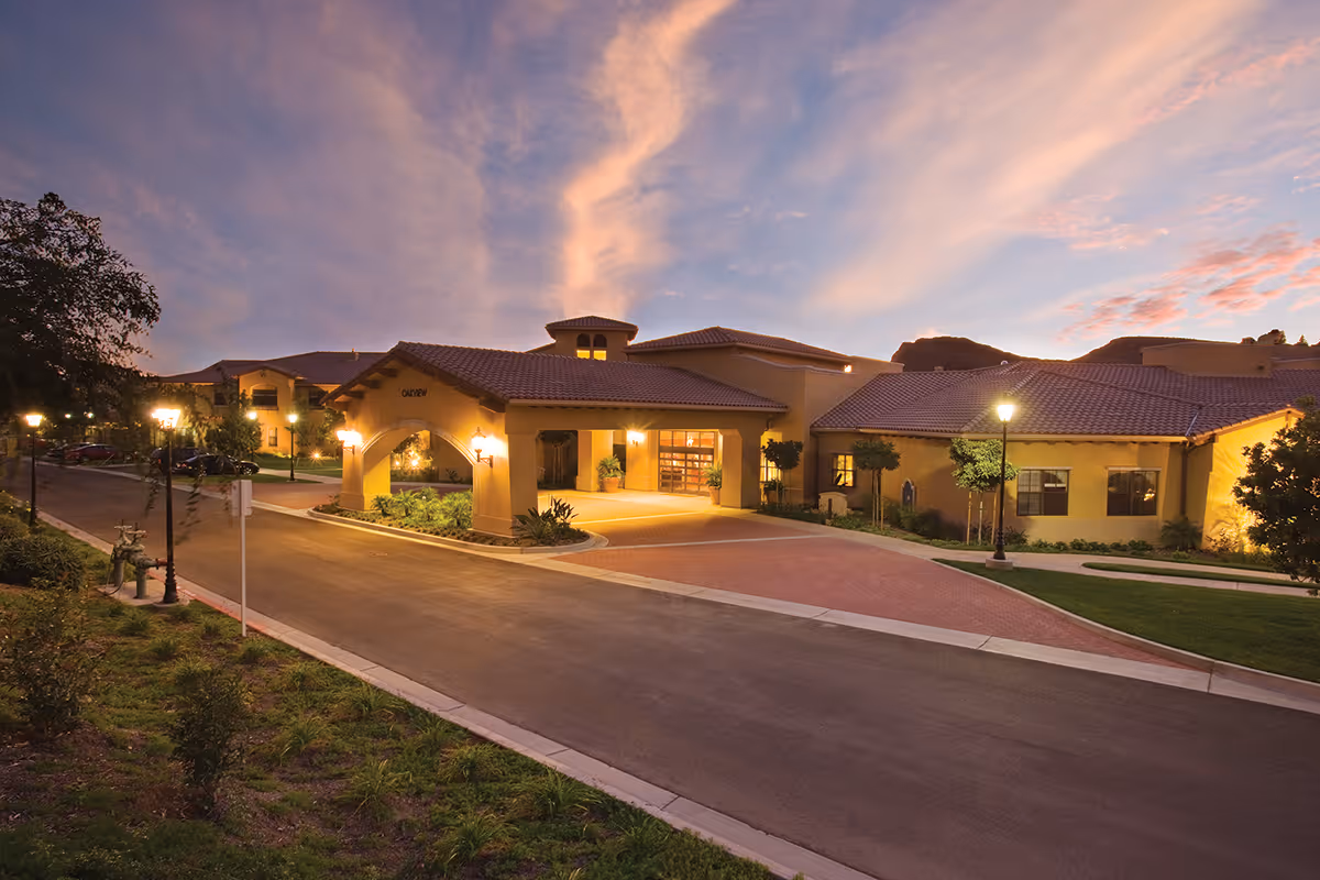 Front entrance and driveway of a landscaped senior living facility at dusk with warm exterior lighting.