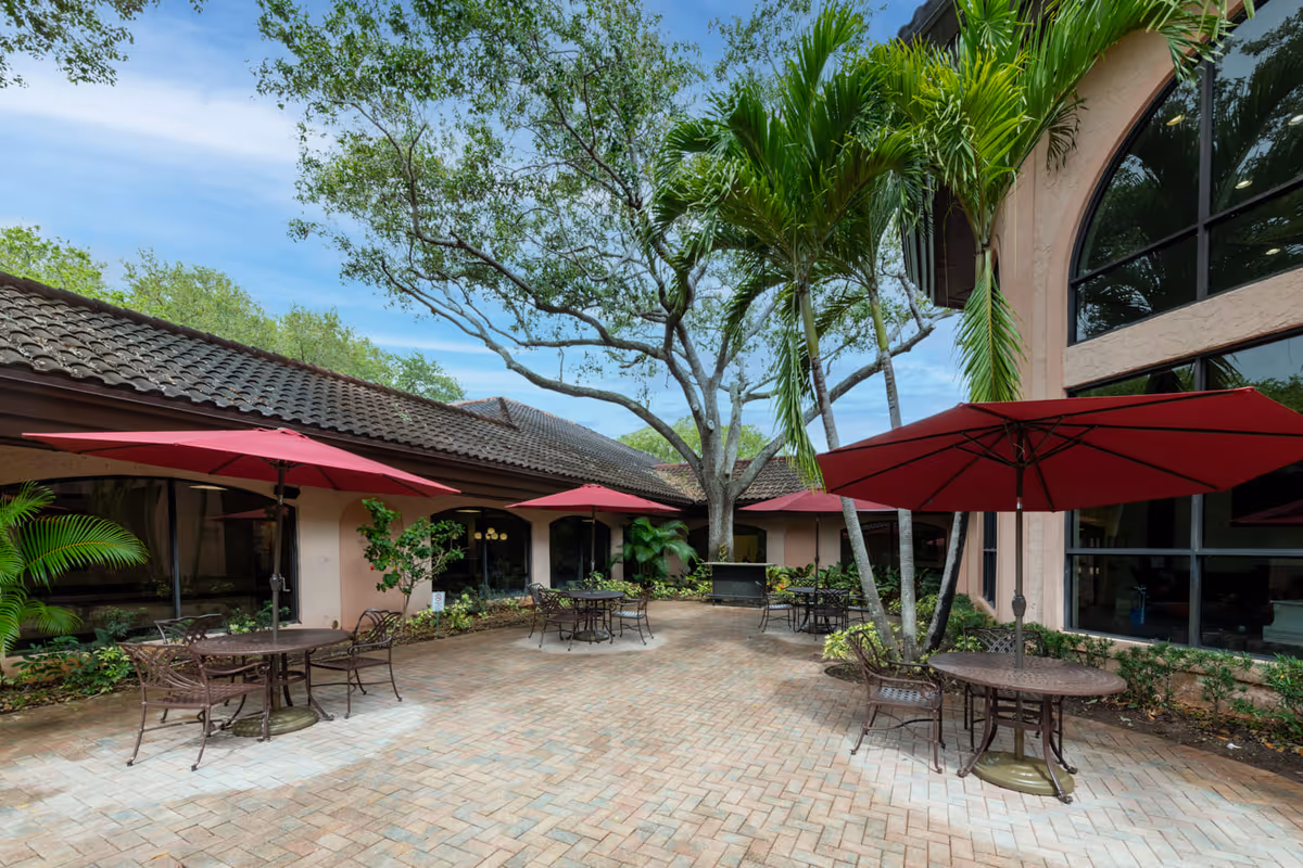 Outdoor courtyard area with brick paver flooring, several round metal tables with chairs, and large red umbrellas providing shade. The courtyard is surrounded by a building with large windows and tropical palm trees and other greenery.
