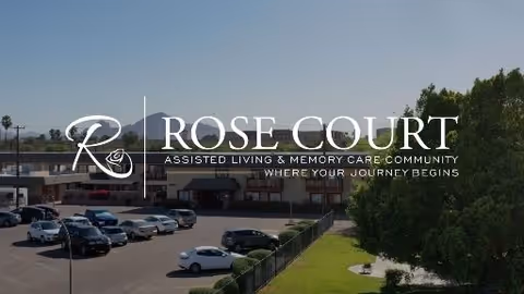 Exterior view of Rose Court Senior Living facility showing a parking lot with several cars, a two-story building in the background, and a grassy area with trees and a fence in the foreground under a clear sky.