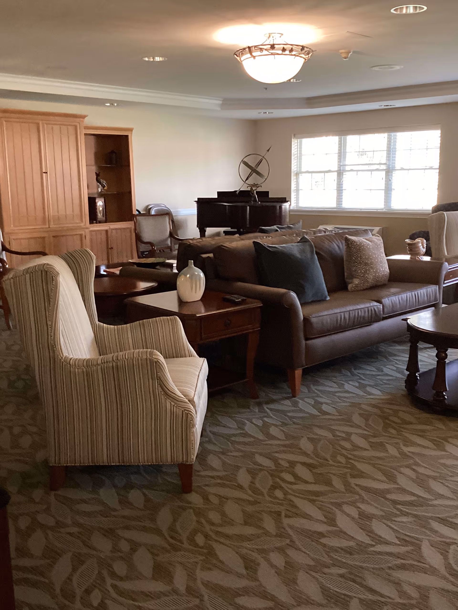 A cozy living room with a brown leather sofa adorned with pillows, a striped armchair, wooden side tables with decorative items, a large window letting in natural light, a wooden cabinet, and a grand piano in the background.