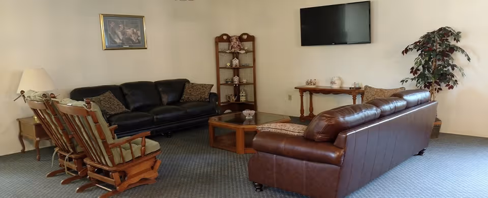 A living room with two leather sofas, two wooden rocking chairs with cushions, a wooden coffee table, a side table with a lamp, a wall-mounted flat screen TV, a decorative shelf with small items, a framed picture on the wall, and a potted plant in the corner.
