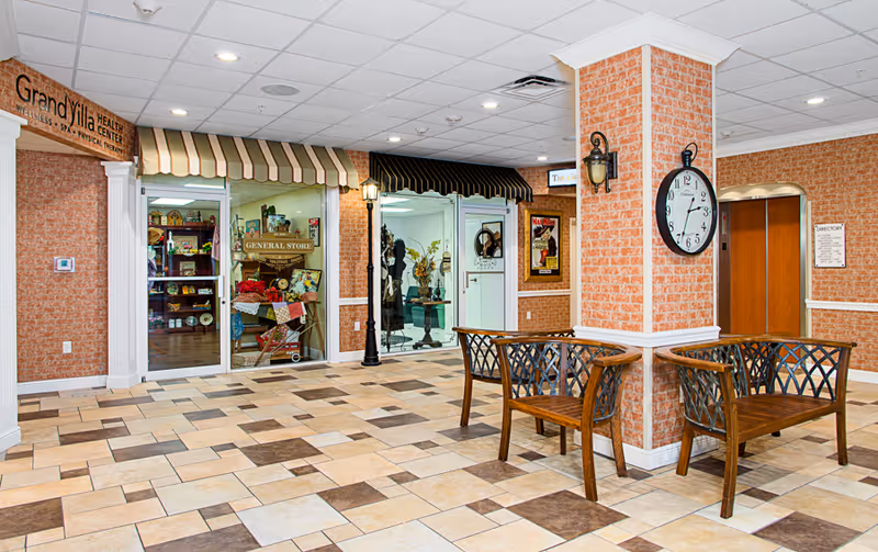 Bright lobby-like interior of a senior living facility with tiled floors, wooden benches around a clock-faced column, and glass-fronted storefronts including a general store.