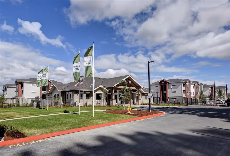 Exterior view of LARC at Kent senior living facility with a clear blue sky and scattered clouds. The image shows a single-story building with a peaked roof, surrounded by well-maintained green lawns and several flagpoles with green and white flags. In the background, there are multi-story residential buildings. The driveway and parking area are visible in the foreground.